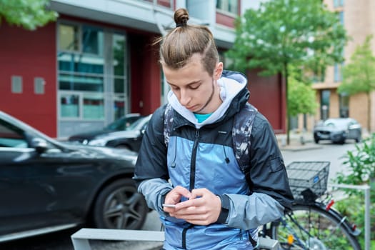 Fashionable young male with backpack using smartphone outdoors in city. Hipster with trendy hairstyle beard with phone in hands mobile applications for tourism travel communication entertainment study