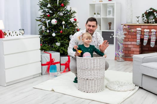 A little girl combed into two ponytails plays in a room. A child of children in a large wicker basket. In the back crouches a man embracing his little daughter and smiling. A decorated Christmas tree can be seen in the background.