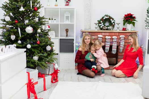 Under a large decorated Christmas tree lie boxes wrapped in red ribbons. Gifts waiting under the Christmas tree. In the background you can see a family of four: Two mothers and two children spending time by the brick fireplace.