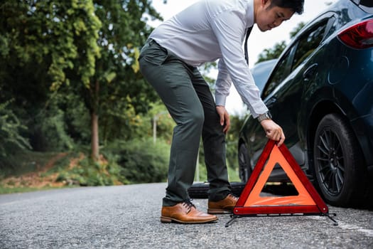 Asian businessman on side of road with broken down red warning triangle. Young male having problem with her car, it stopped on the street. Stranded at Roadside and Waiting for Help.