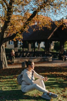 Young fashionable teenage girl with smartphone in park in autumn sitting at smiling. Trendy young woman in fall in park texting. Retouched, vibrant colors. Beautiful blonde teenage girl wearing casual modern autumn outfit sitting in park in autumn. Retouched, vibrant colors, brownish tones.