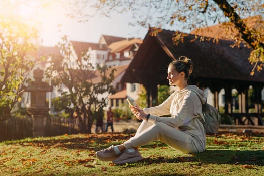 Young fashionable teenage girl with smartphone in park in autumn sitting at smiling. Trendy young woman in fall in park texting. Retouched, vibrant colors. Beautiful blonde teenage girl wearing casual modern autumn outfit sitting in park in autumn. Retouched, vibrant colors, brownish tones.