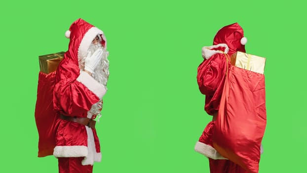 Romantic man in santa costume giving air kisses, cute while he is carrying traditional festive sack with gifts. Sweet saint nick does flirty gesture standing over greenscreen backdrop.
