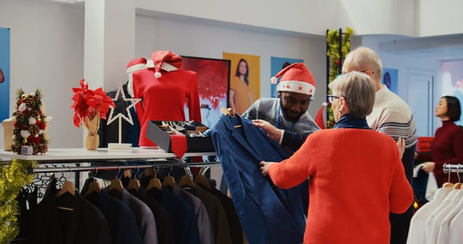 Wife and husband in xmas ornate shopping mall fashion shop, being assisted by african american worker with finding ideal outfit during festive holiday season promotional sales