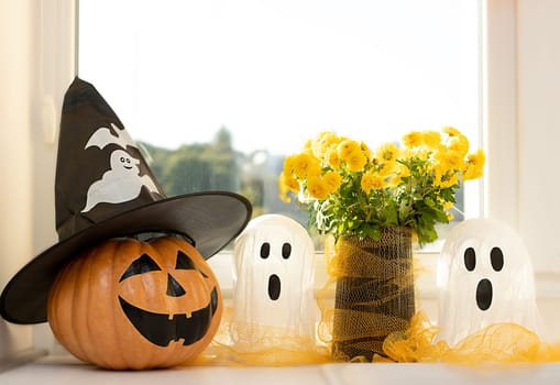 Halloween concept. A pumpkin with a painted face, a white ghost and a bouquet of yellow chrysanthemum flowers in a black vase against the background of a window in home interior. Close-up