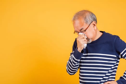 Portrait senior old man blowing nose with tissue studio shot isolated on yellow background, Asian elder man cold having flu and sneezing from sickness virus problem use tissue, pensioner unwell