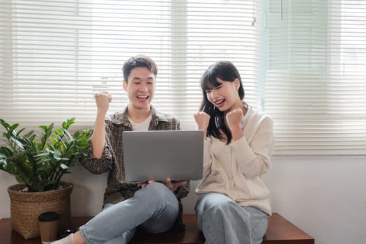 A group of cheerful young university students are looking at a laptop screen and showing their fists, celebrating a good news together while sitting in a cafe..