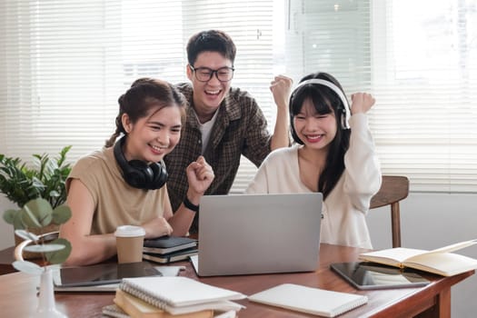 A group of cheerful young university students are looking at a laptop screen and showing their fists, celebrating a good news together while sitting in a cafe..