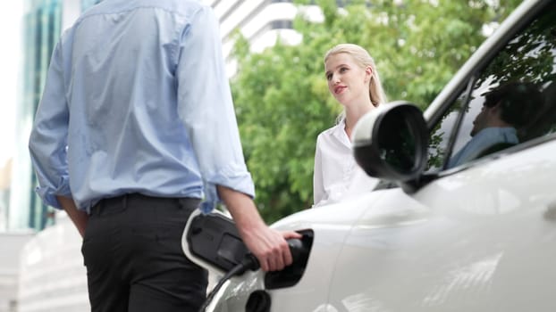 Progressive businessman and businesswoman install charger plug from charging station to electric car before driving around city center. Eco friendly rechargeable car powered by clean energy.