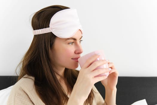 Portrait of beautiful woman relaxing in bed, waking up and having her morning cup of coffee in bedroom, looking happy.