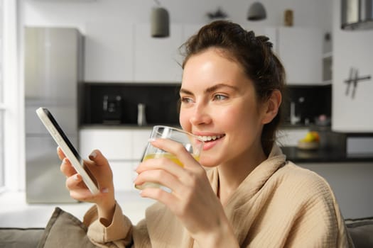 Close up portrait of laughing, smiling young woman enjoys morning, drinking orange juice and holding smartphone, reading news on mobile app.