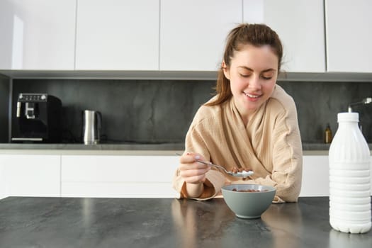 Portrait of happy young woman leans on kitchen worktop and eating cereals, has milk and bowl in front of her, having her breakfast, wearing bathrobe.