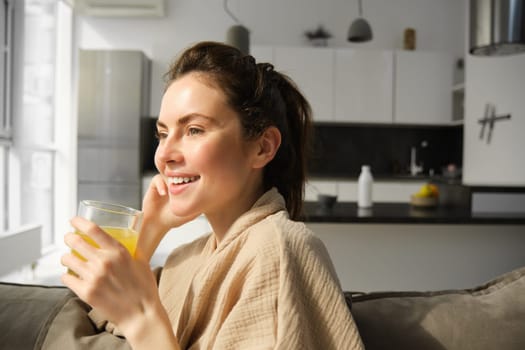 Close up of beautiful young woman on sofa, drinks orange juice and smiles, looks outside window, enjoys morning at home.