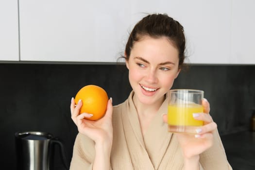 Image of good-looking healthy woman in bathrobe, drinking fresh juice, showing orange fruit, posing in kitchen.