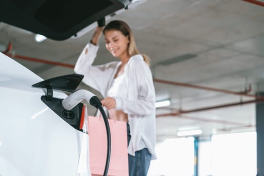 Young woman travel with EV electric car to shopping center parking lot charging in downtown city showing urban sustainability lifestyle by green clean rechargeable energy of electric vehicle innards