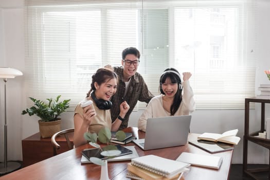 A group of cheerful young university students are looking at a laptop screen and showing their fists, celebrating a good news together while sitting in a cafe..