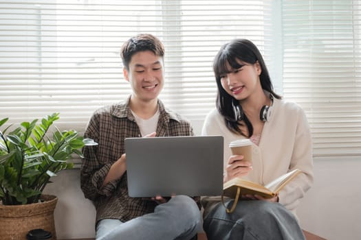 A happy young Asian man and a pretty girl are working on a laptop together, working on a co-project, sharing ideas and discussing work in a room..