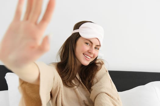 Hotel and vacation. Young woman waking up in her room and smiling, has sleeping mask on forehead, stretching her arm towards camera and smiling, resting, waking up in morning.