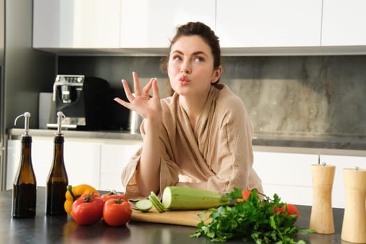 Portrait of beautiful young woman in bathrobe, cooking meal for family, shows chefs kiss, okay sign, making food, preparing vegetarian dinner, chopping vegetables.