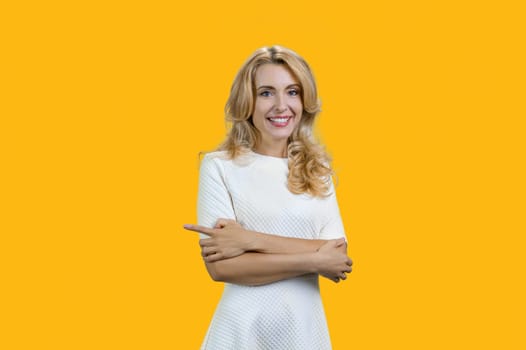 Portrait of happy smiling blonde woman with folded arms pointing to the right. Isolated on yellow.