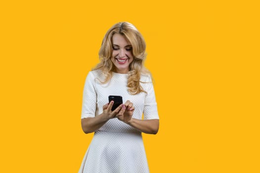 Happy blonde mature woman using mobile phone. Typing sms message. Isolated on yellow.