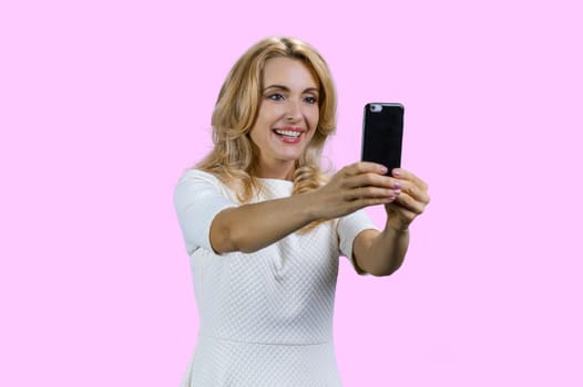 Portrait of happy blonde woman taking selfie on smartphone frontal camera. Isolated on pink.