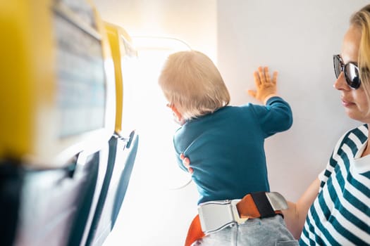 Mom and child flying by plane. Mother holding and playing with her infant baby boy child in her lap during economy comercial flight. Concept photo of air travel with baby. Real people