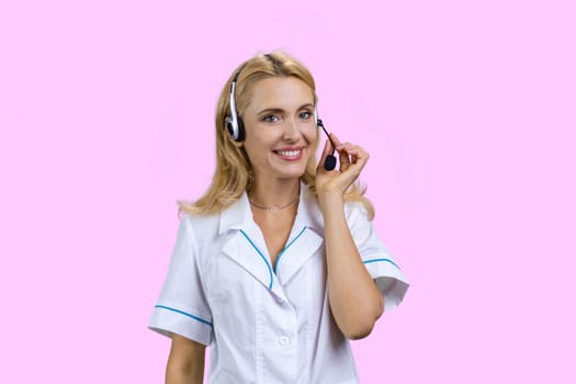 Portrait of cheerful blonde mature female doctor wearing headset. Isolated on pink.