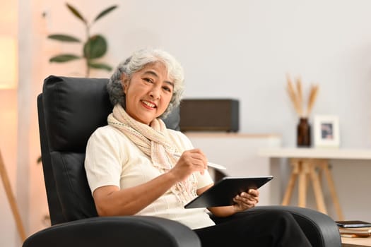 Beautiful senior woman sitting on comfortable armchair and using digital tablet. Elderly lifestyle concept.