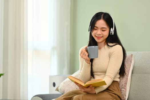 Carefree young woman in headphone drinking coffee and reading book on couch.