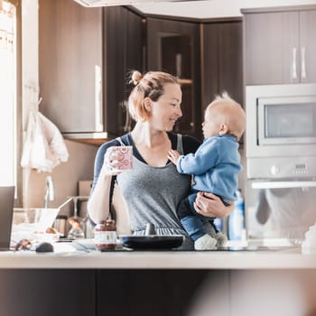 Happy mother and little infant baby boy together making pancakes for breakfast in domestic kitchen. Family, lifestyle, domestic life, food, healthy eating and people concept