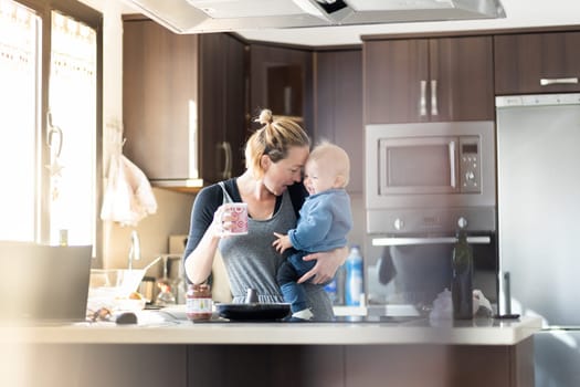 Happy mother and little infant baby boy together making pancakes for breakfast in domestic kitchen. Family, lifestyle, domestic life, food, healthy eating and people concept