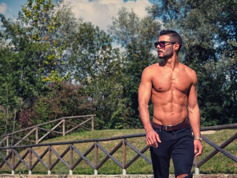 A shirtless handsome athletic man walking down a path in a park.