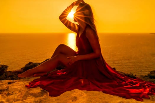 Woman sunset sea red dress, side view a happy beautiful sensual woman in a red long dress posing on a rock high above the sea on sunset