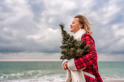 Blond woman holding Christmas tree by the sea. Christmas portrait of a happy woman walking along the beach and holding a Christmas tree in her hands. Dressed in a red coat, white dress