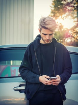 A man standing in front of a car looking at his cell phone. Photo of a man using his cell phone while standing in front of a car