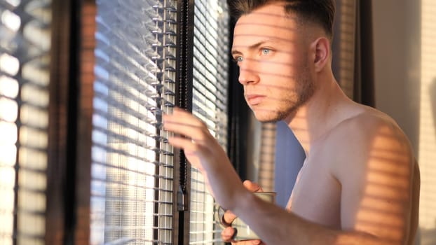 A shirtless man looking out of a window. Photo of a bare-chested man peering out of a window