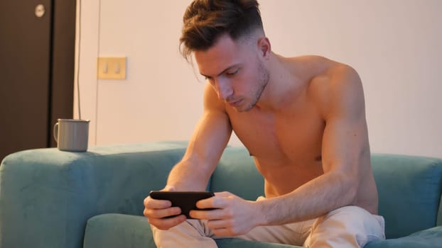 A shirtless man sitting on a couch looking at a cell phone. Photo of a shirtless man using a smartphone on a couch