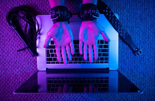 the female sexy hands in bdsm handcuffs with laptop in neon light on dark background