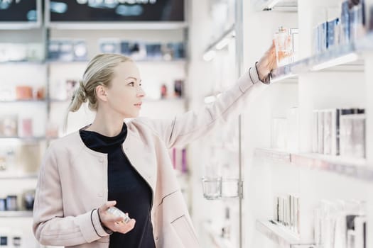 Elegant blond young woman choosing perfume in retail store. Beautiful blond lady testing and buying cosmetics in a beauty store.