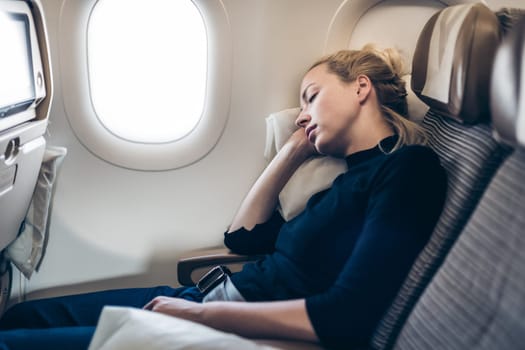 Tired blonde casual caucasian lady napping on seat while traveling by airplane. Commercial transportation by planes.