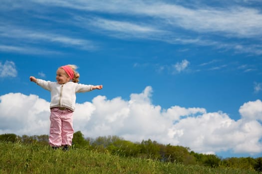 A little girl cheers with her arms raised in a meadow against a blue,slightly cloudy sky