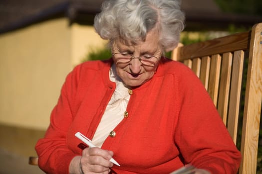 An elderly lady sits on a chair and writes something down or solves a riddle