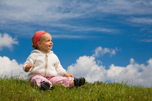A little girl sits on a summer meadow against a blue slightly cloudy background