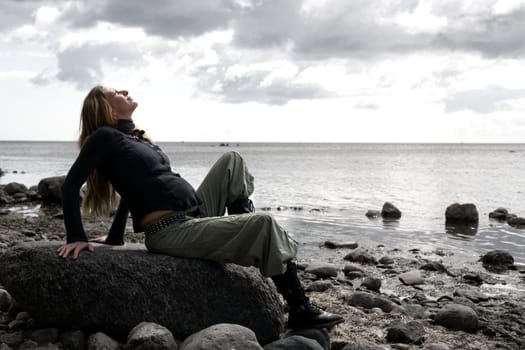 A young pregnant woman sits on a stone on the beach and looks up at the sky
