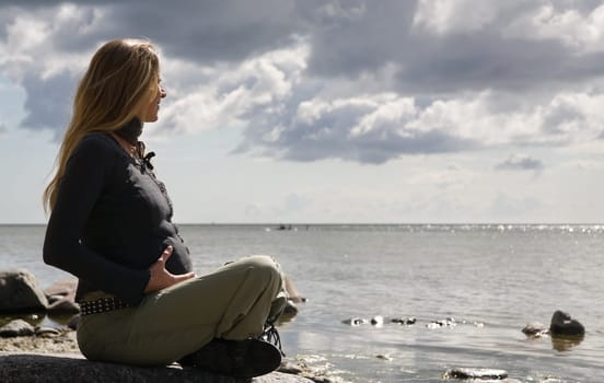 A young pregnant woman sits on a stone on the beach,holds her stomach and looks relaxed into the distance