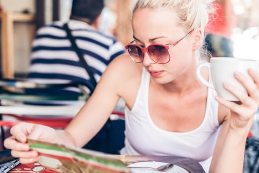 Calm casual blond lady wearing fashionable sunglasses, enjoying cup of coffee while checking menu in typical italian street coffee house or restaurant on warm summer day.