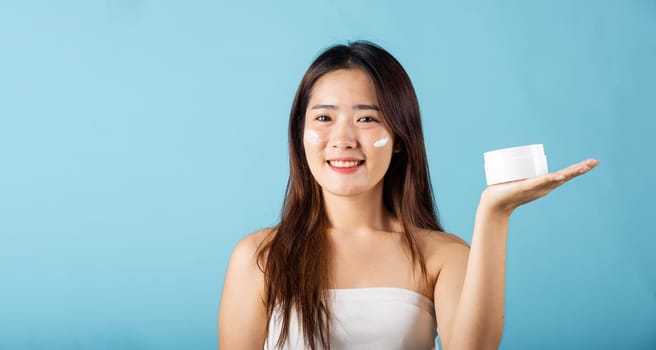 Asian young woman are not very beautiful hold jar of skin cream for face and apply anti-aging moisturizer studio isolated on blue background, Happy female pointing finger her face, pursuit of beauty