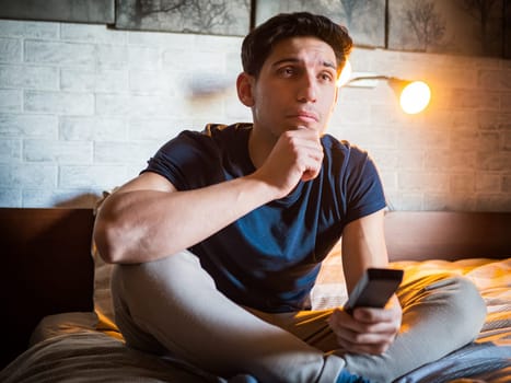 Attractive young man laying on his bed holding remote control, watching TV with uneasy, worried expression