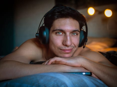 Attractive Young Man Using Headphones Lying Alone On His Bed, Listening to Music and Having a Good Time, Looking at Camera